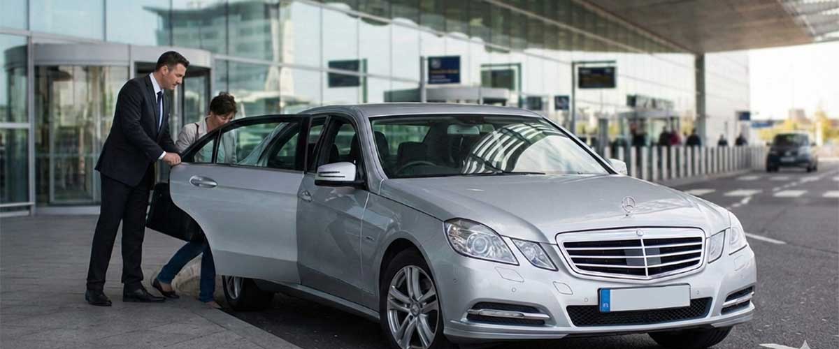 Premium airport transfer service in Ashford Kent. A professional Ashford Infinity Cabs driver in a suit holding the door open for a passenger entering a silver Mercedes executive car at an airport terminal.