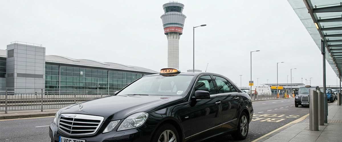 Ashford Infinity Cabs black Mercedes taxi parked outside London Heathrow Airport Terminal 5, with the iconic control tower in the background. Professional airport transfer service.